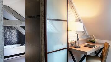 A modern bedroom with a sloped ceiling, a bedside table with a lamp, a chair, and a small desk with a laptop.