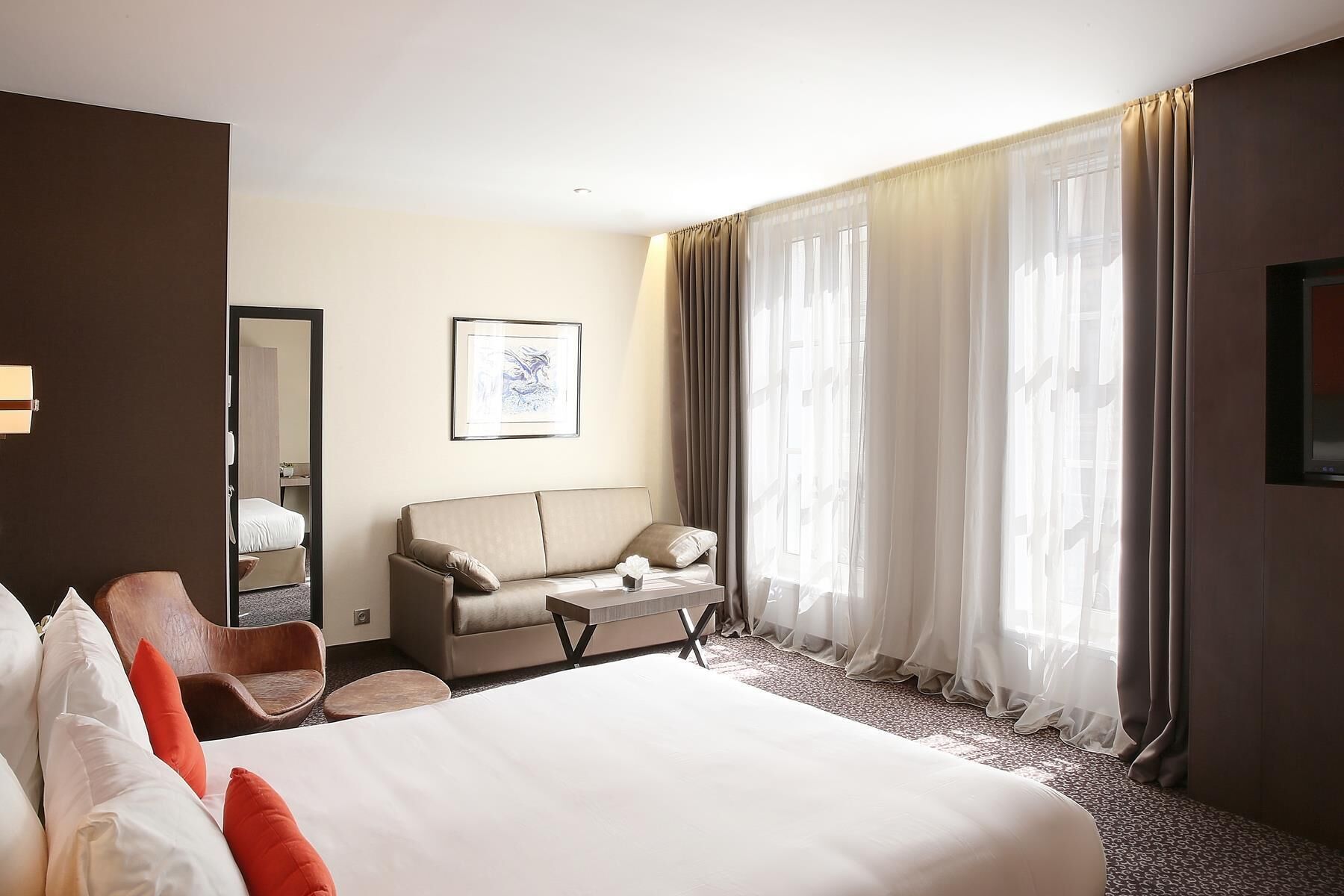 A hotel room with a large bed, a sofa, a chair, a desk, a mirror, and sheer curtains.