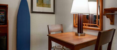 A hotel room with a wooden table, two chairs, a blue ironing board, a lamp, and a framed artwork on the wall.