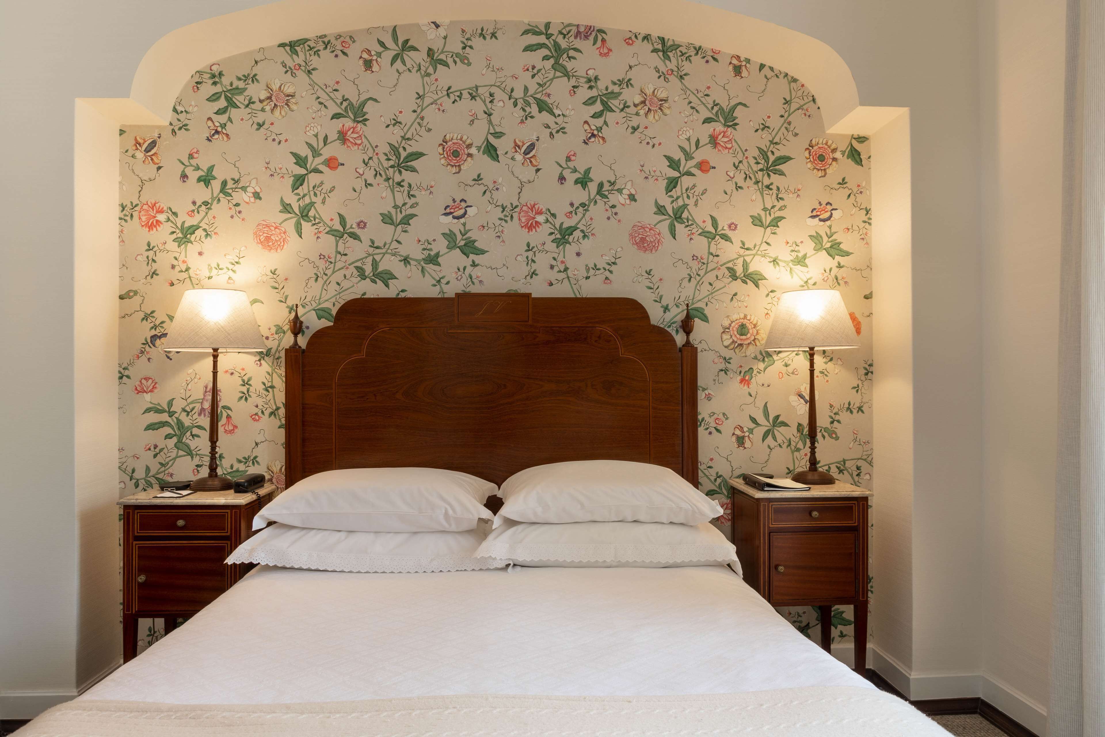 A bedroom with a floral wallpaper, a wooden headboard, two bedside tables with lamps, and a bed with white linens.