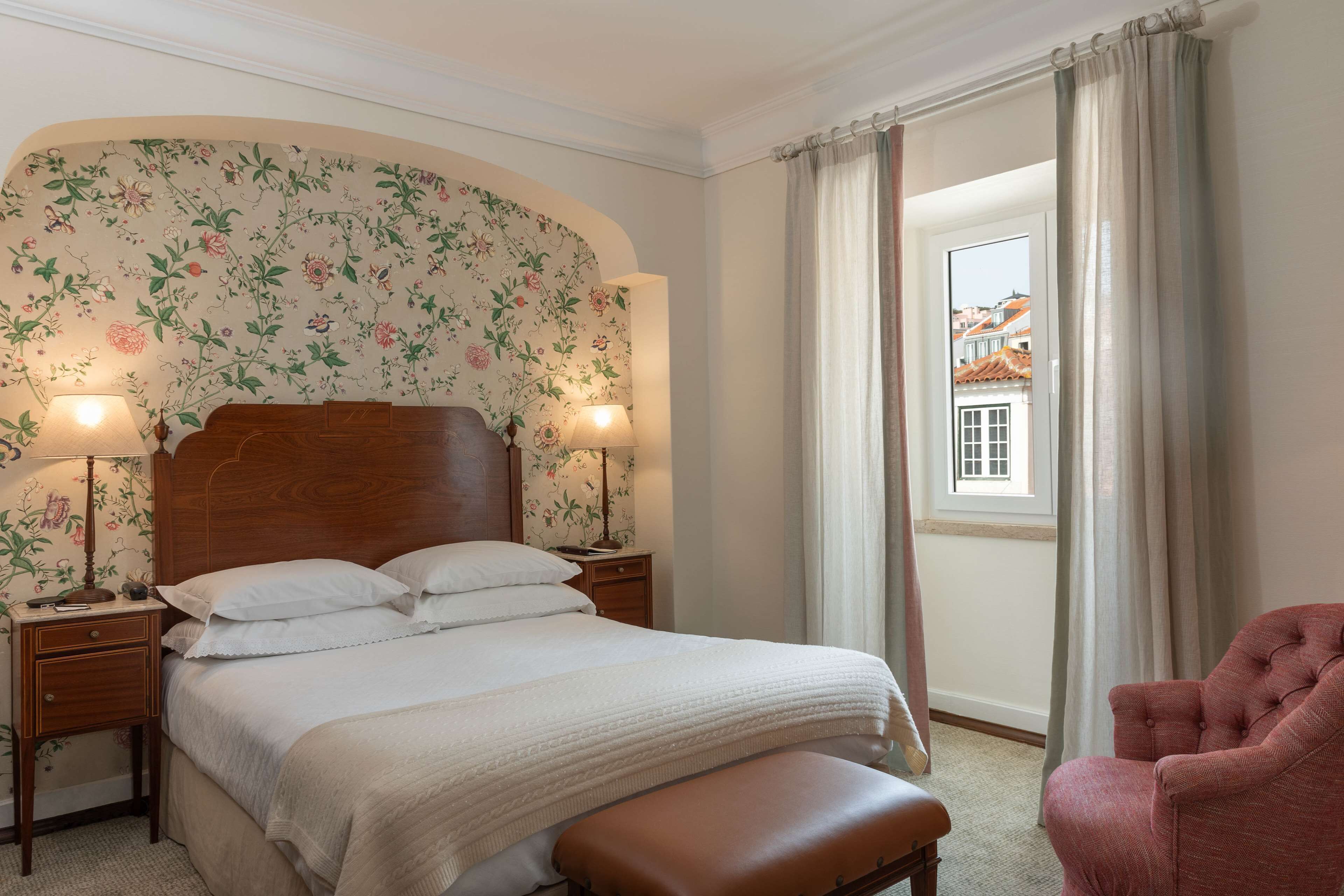 A neatly made bed with a wooden headboard, two bedside lamps, a small bedside table, and a window with curtains.