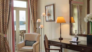 Een hotelkamer met een bed, twee fauteuils, een bureau met een lamp, een spiegel en uitzicht op zee door het raam.