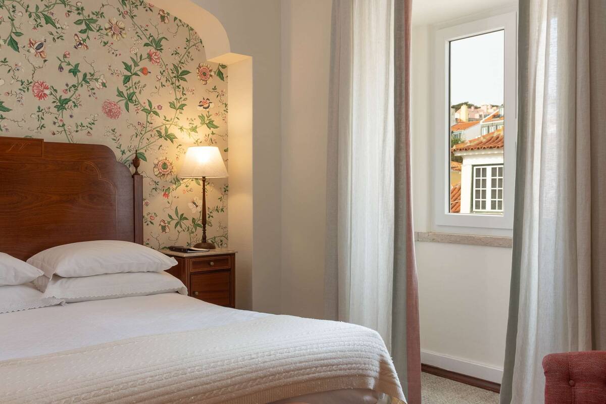 Una habitación de hotel con cama, un banco, una silla, una mesita de noche con lámpara y una ventana con cortinas.