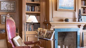 A library with bookshelves, a fireplace, a reading chair, and a desk with a lamp.