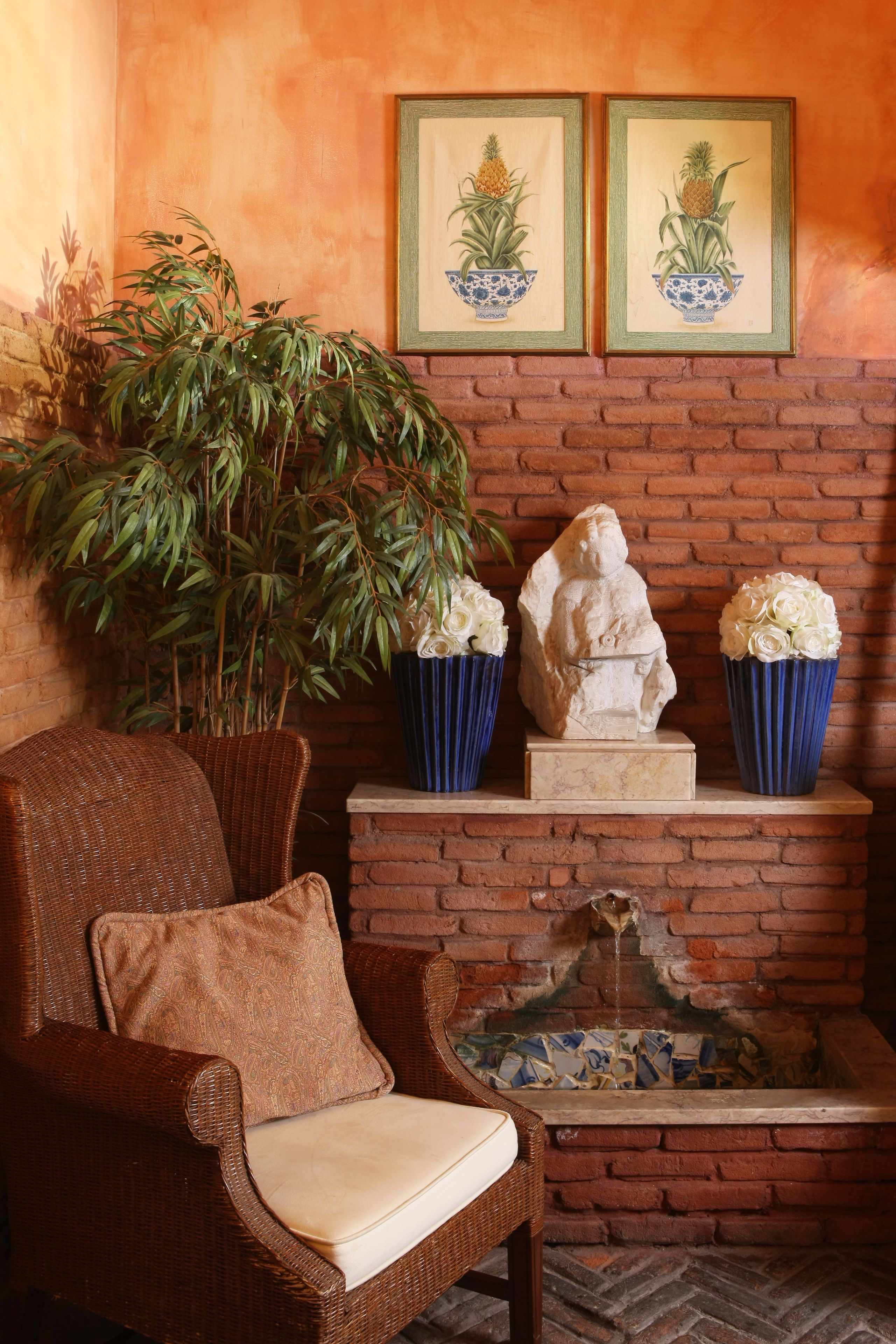 Une chaise en osier avec un coussin, une grande plante en bambou, une sculpture en pierre et deux tableaux encadrés représentant des ananas, le tout placé contre un mur de briques.