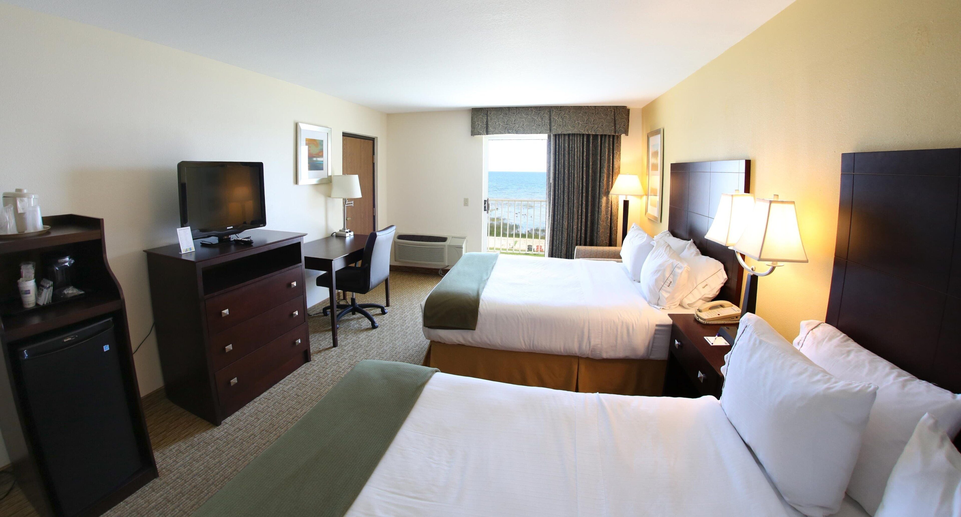 A hotel room with two beds, a desk, a television, a mini-fridge, and a view of the ocean.