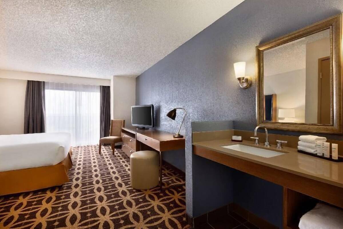 Een hotelkamer met een bed, een bureau met een televisie, een grote spiegel, een wastafel en een kamerbreed tapijt met dessin.