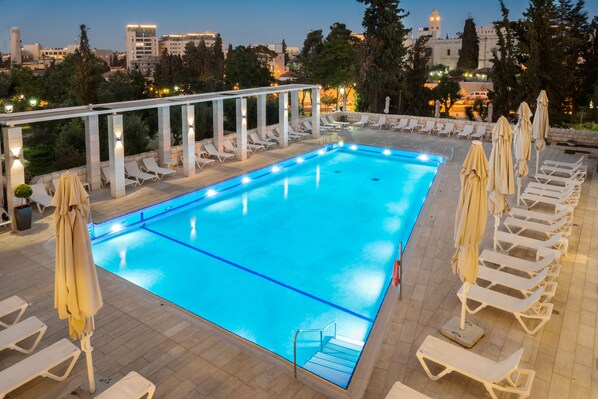 Una piscina rectangular con superficie de agua azul cristalina, rodeada de sillones blancos y sombrillas.