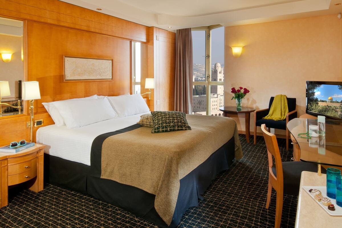 A hotel room with a large bed, a desk with a computer, a chair, a small table with a vase of flowers, and a window with a city view.