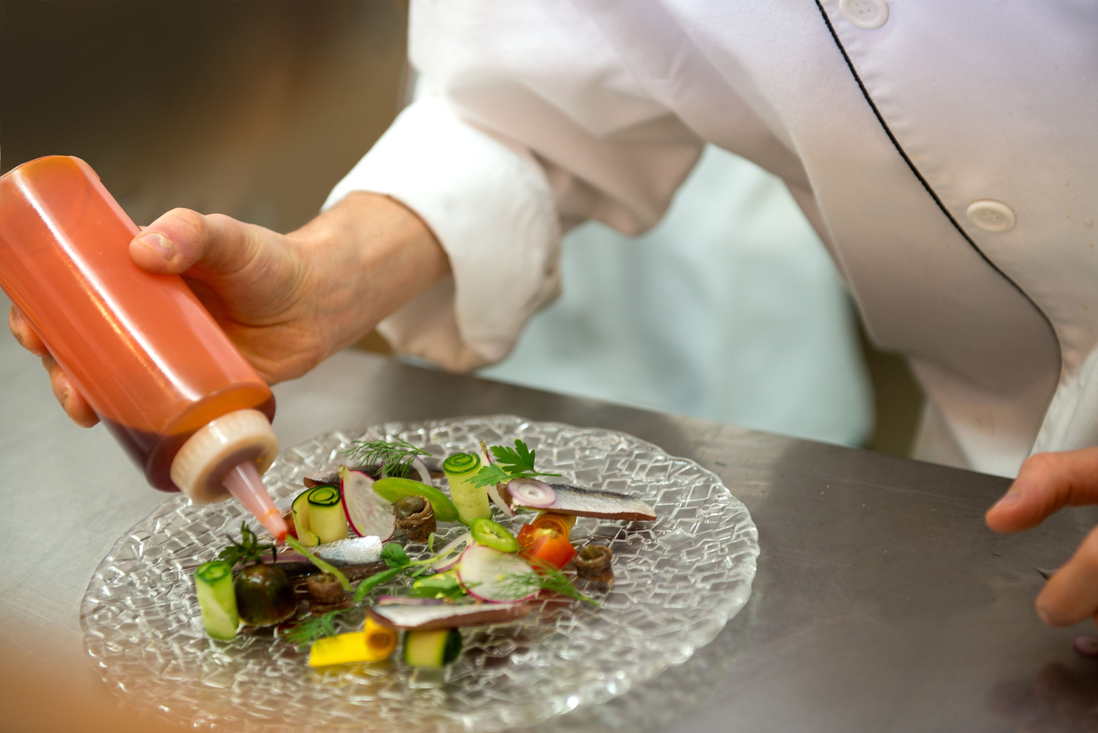A chef preparing a dish with a variety of vegetables and fish on a glass plate.