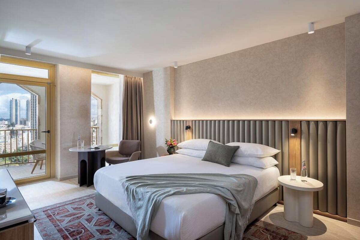 A modern hotel room with a large bed, a small desk, a round side table, and a view of the city through a large window.
