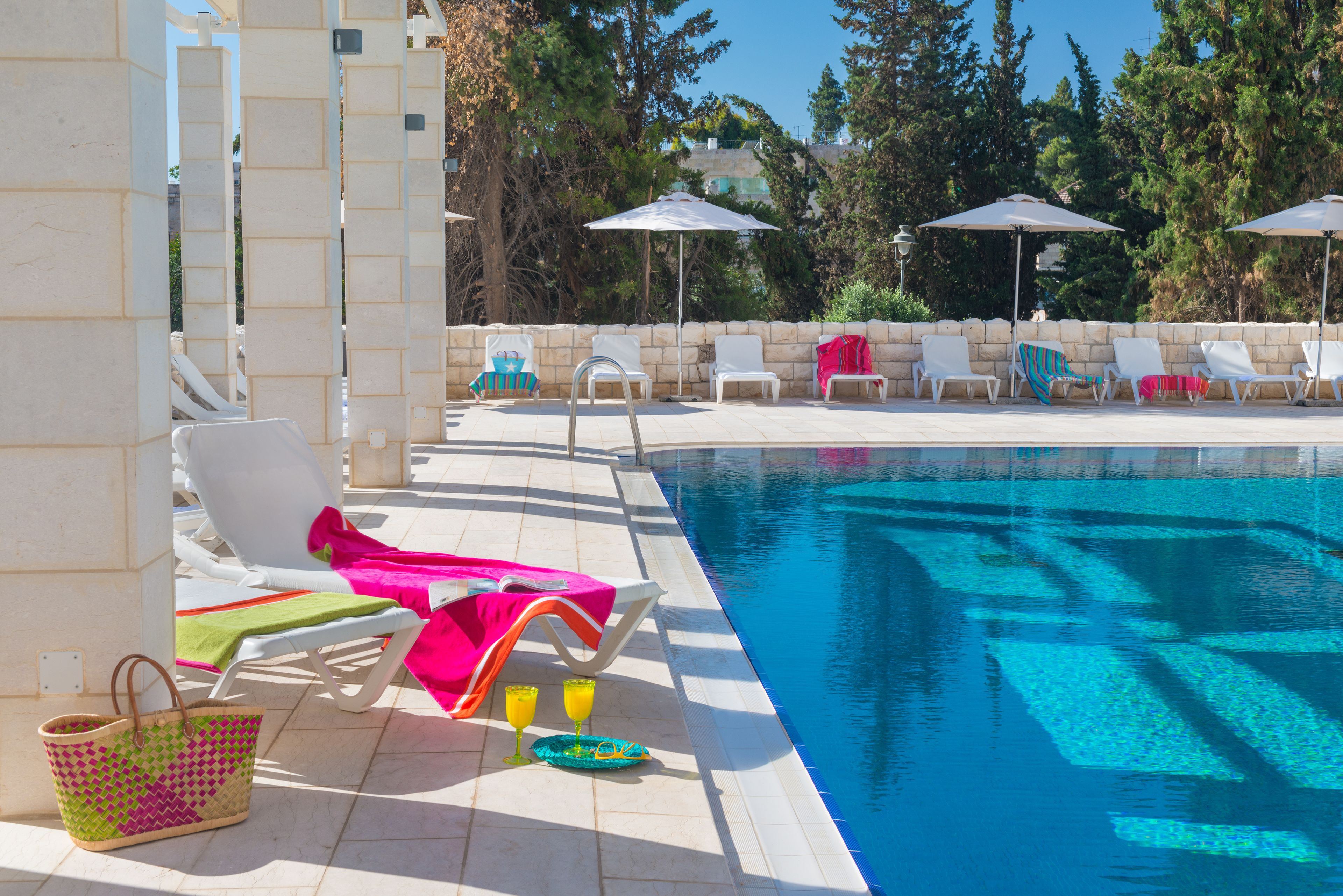 Un área junto a la piscina con sillones de descanso y sombrillas, una piscina cristalina y un muro de piedra.