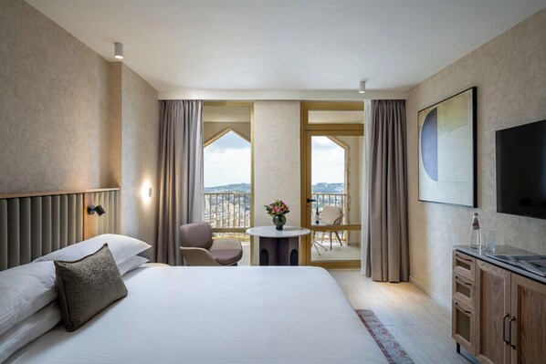 A hotel room with a bed, a TV, a desk, a chair, a small table with a vase, and a balcony with curtains.