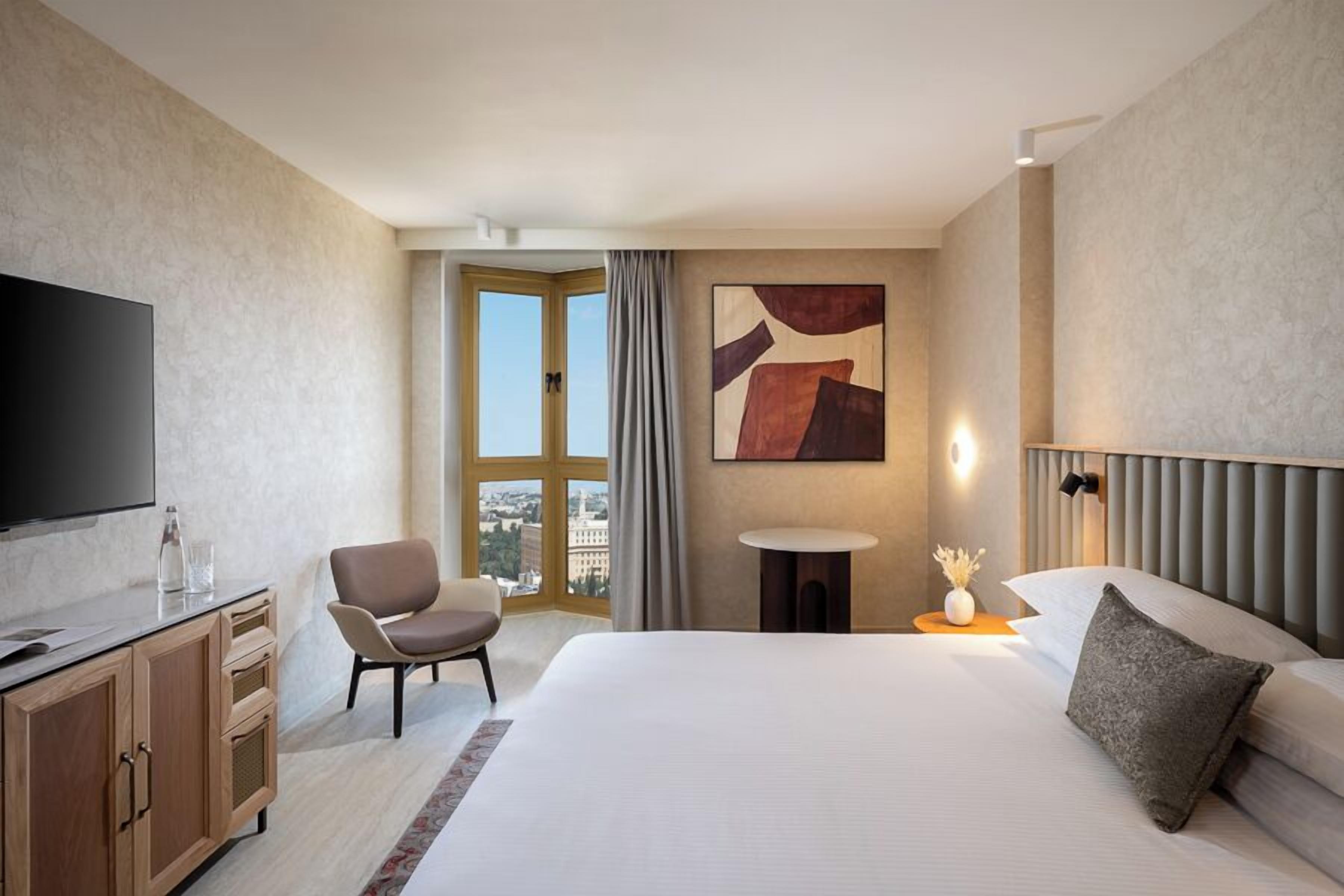 A hotel room with a large bed, a television, a desk, a chair, a window with a view, and a painting on the wall.