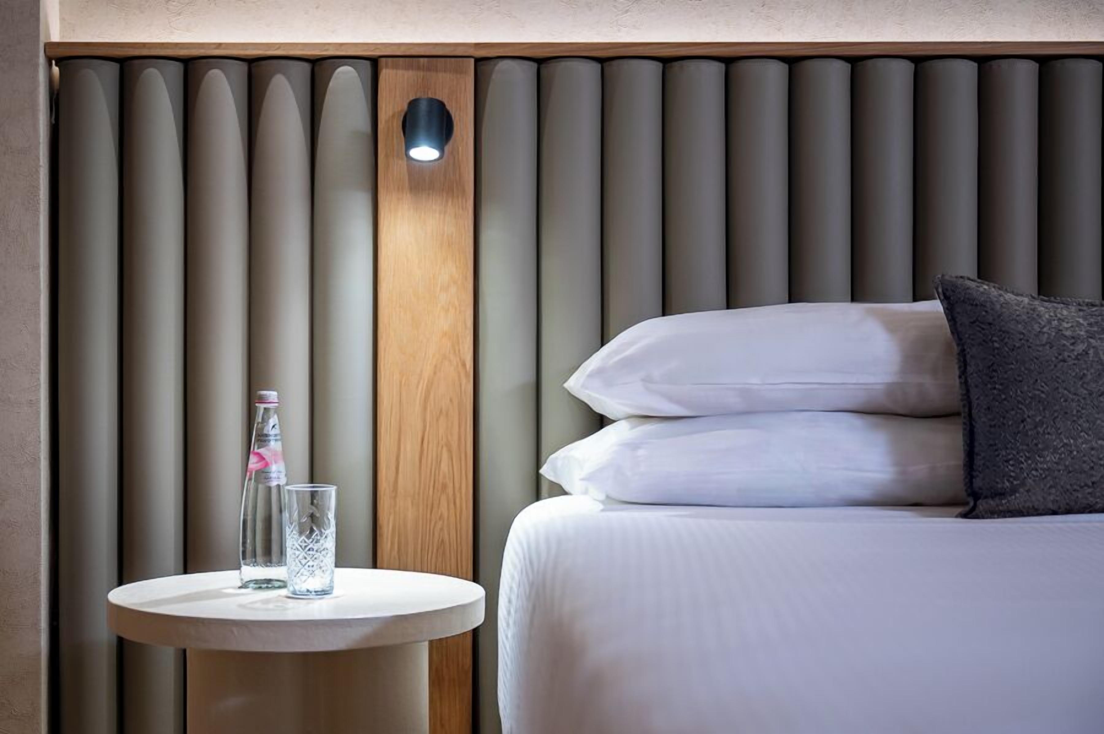 A hotel room with a bed, a round table with a bottle and glass, and a wall with vertical panels.