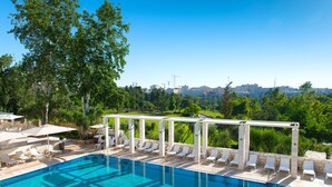 Una piscina con sillas de descanso y sombrillas, rodeada de vegetación y un paisaje urbano de fondo.