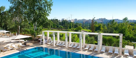 A swimming pool with lounge chairs and umbrellas, surrounded by greenery and a cityscape in the background.