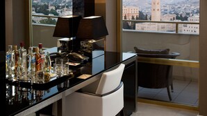 Habitación de hotel moderna con balcón, zona de bar con botellas y vasos, y vista a un paisaje urbano.