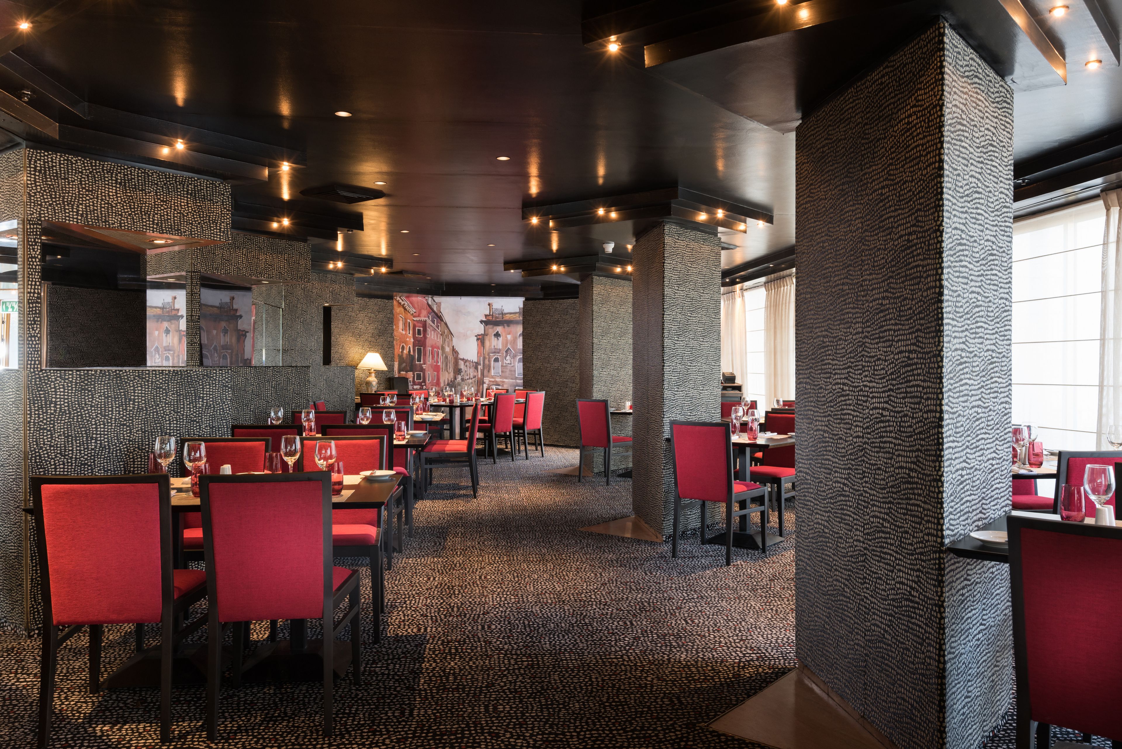 A restaurant with red chairs, tables, and a patterned carpet.
