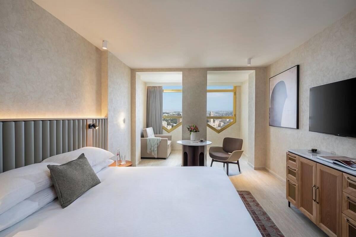A hotel room with a large bed, a TV, a dining area, and a view of the city.