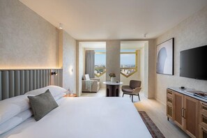 Habitación de hotel con una cama grande, televisión, zona de comedor y vistas a la ciudad.