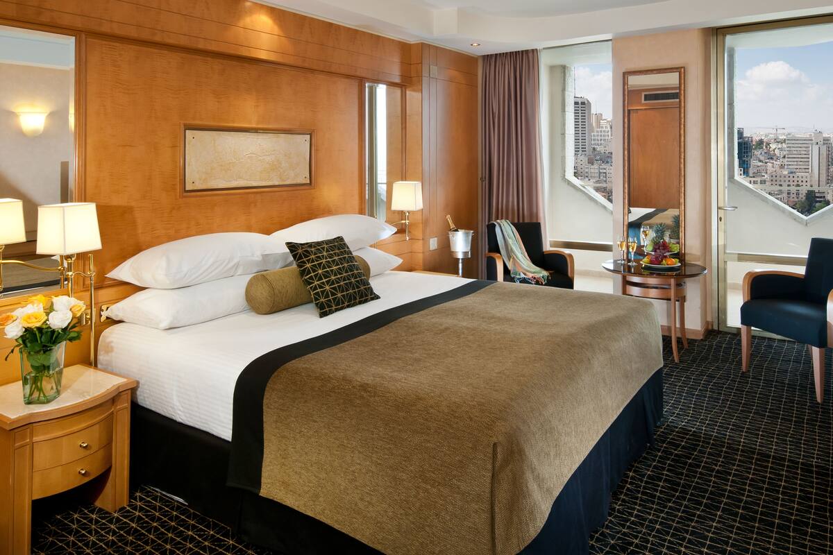 A hotel room with a large bed, a nightstand with a vase of flowers, a chair, a small table with a fruit bowl and drinks, and a city view through large windows.