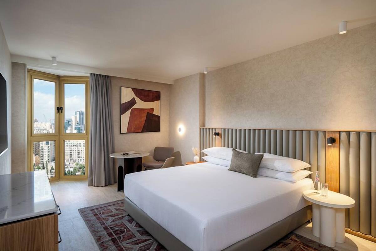 A hotel room with a large bed, a desk, a chair, a small table, and a view of the city through a large window.