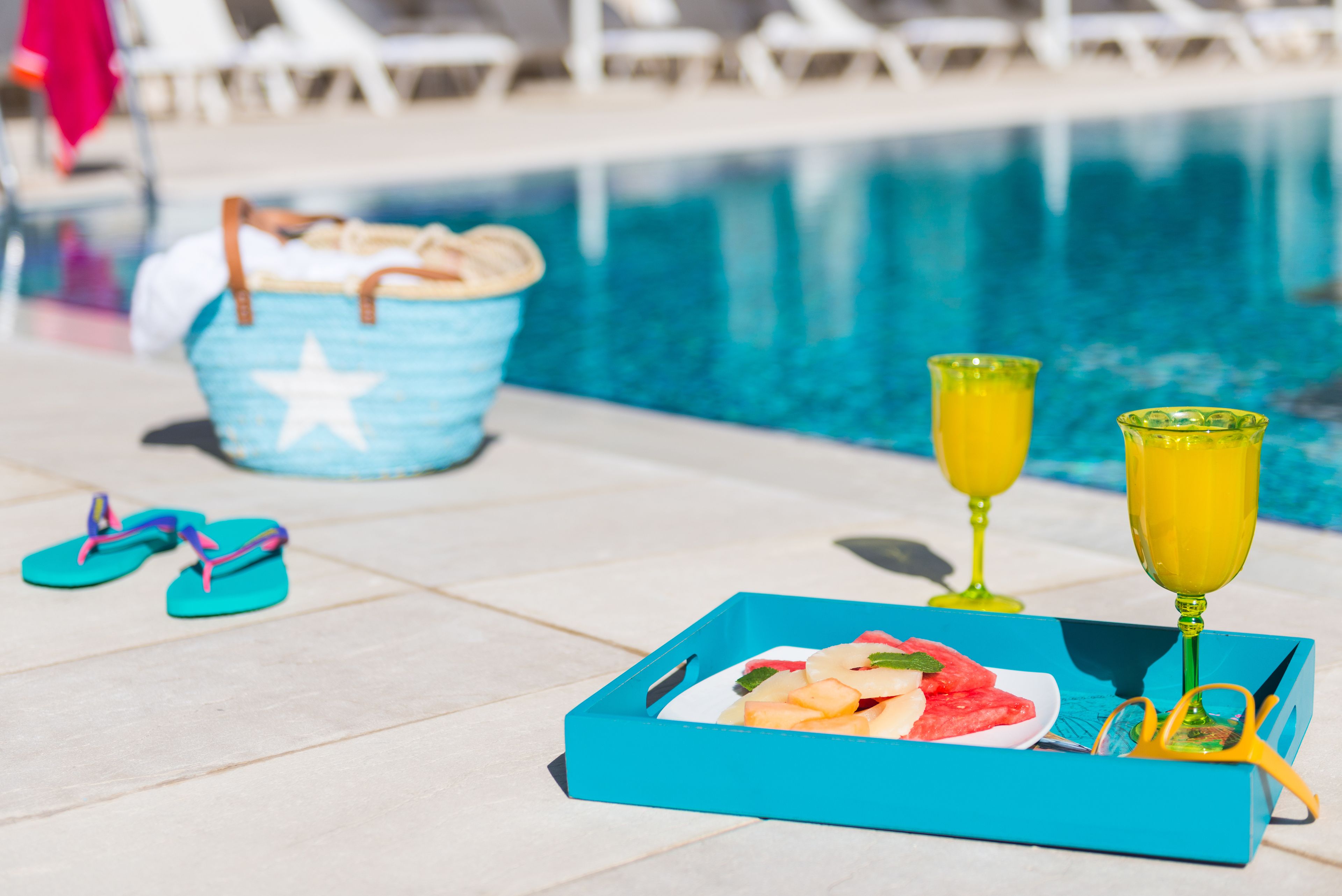 A poolside setting with a tray of fruit, two glasses of orange juice, a pair of flip-flops, and a blue bucket with a star design.