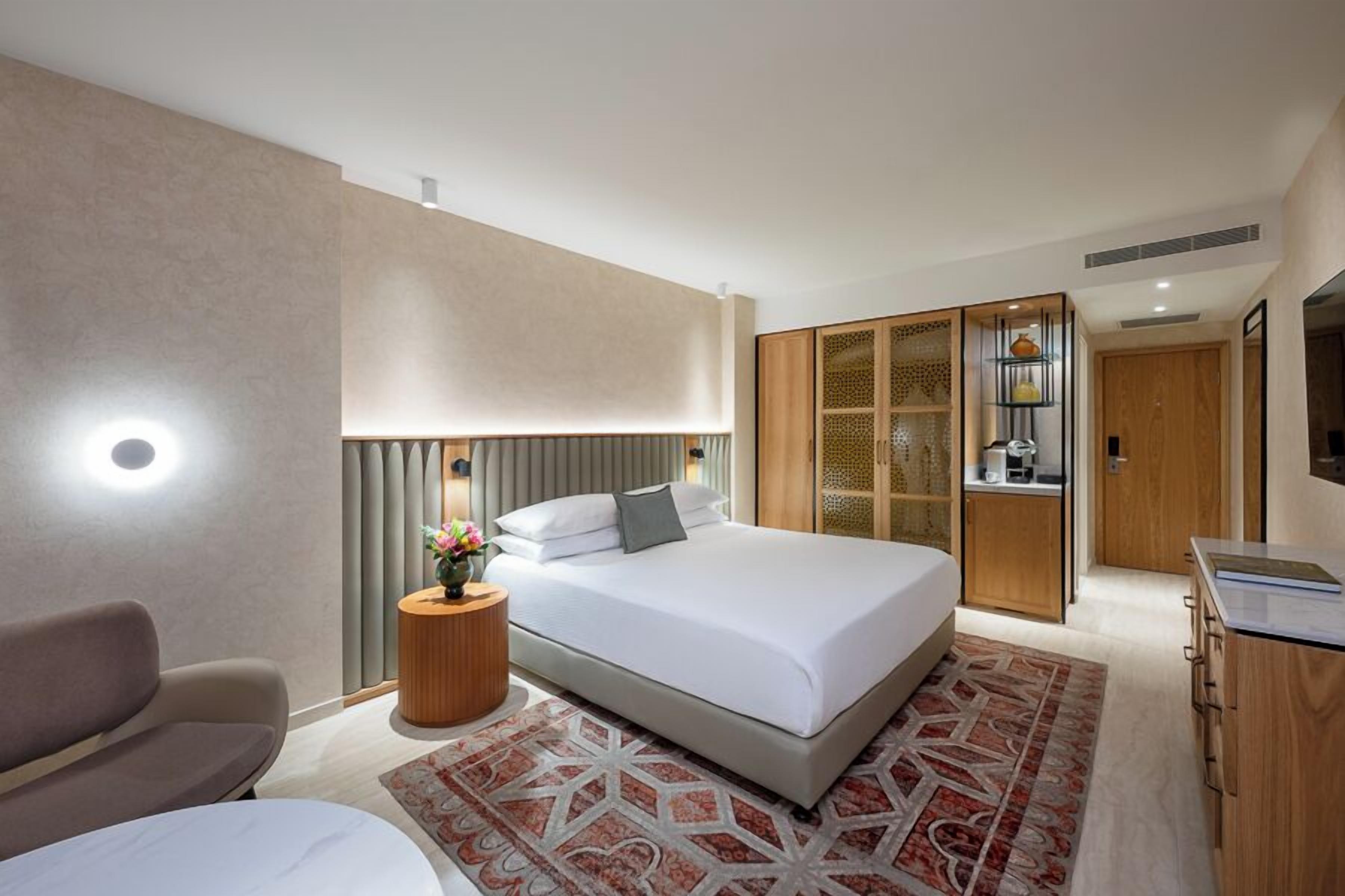 A modern hotel room with a large bed, a sofa, a small side table with flowers, a wooden wardrobe, and a desk with a chair.