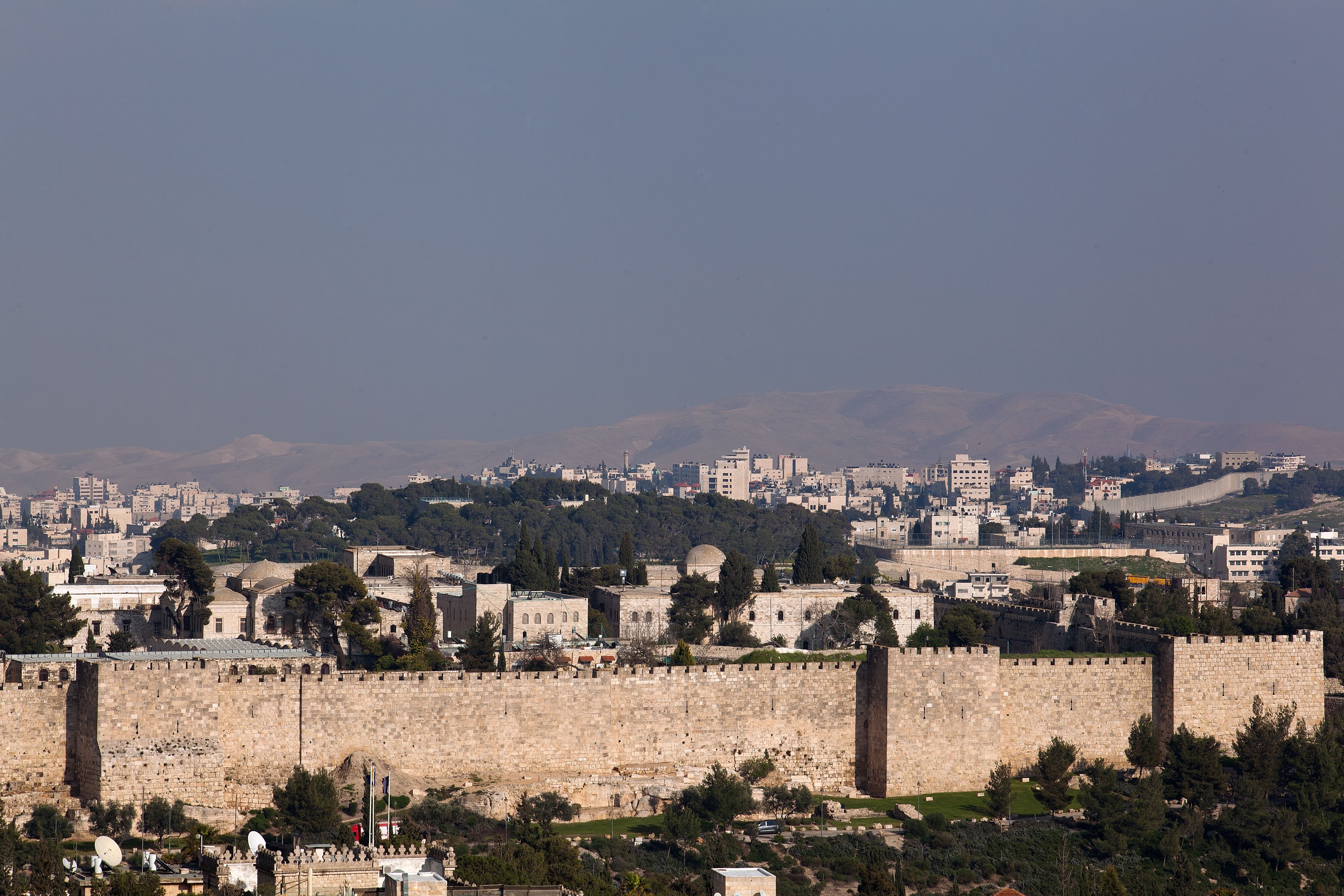 A historic stone wall with a fortified structure, surrounded by greenery and urban buildings in the background.