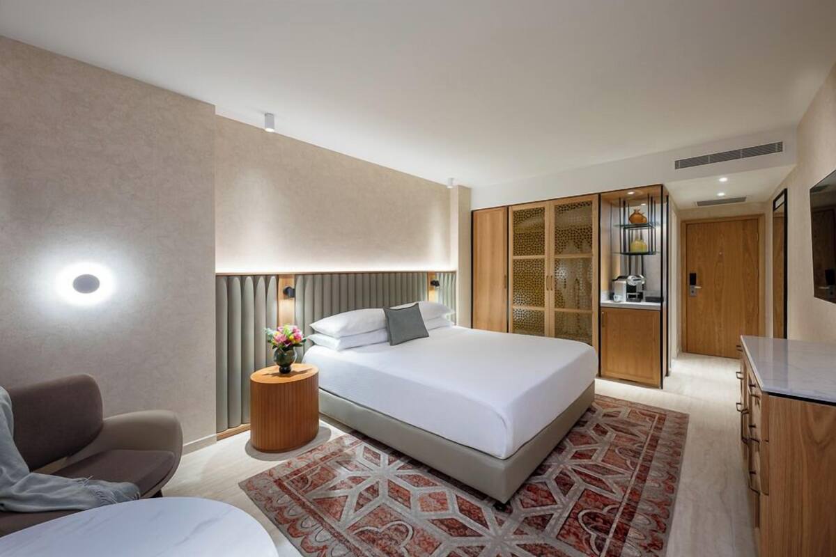 A modern hotel room with a large bed, a seating area, a small table with a vase of flowers, and a wardrobe with a mirror.