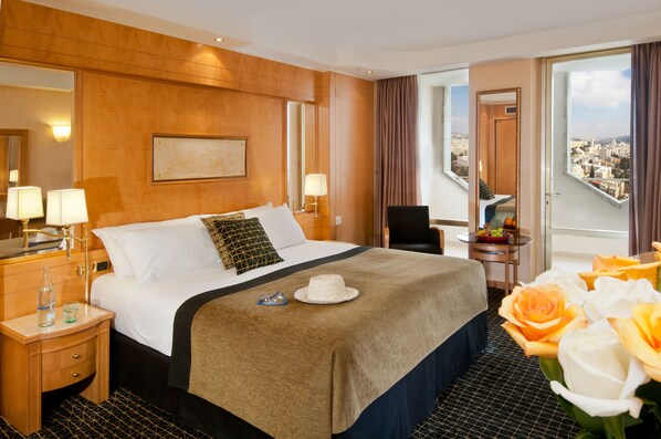 Habitación de hotel con una cama grande, un escritorio, una silla, un ventanal con vista a la ciudad y un balcón con flores.
