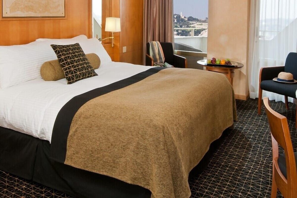 A hotel room with a large bed, a small table with fruit, a chair, and a view of a cityscape.