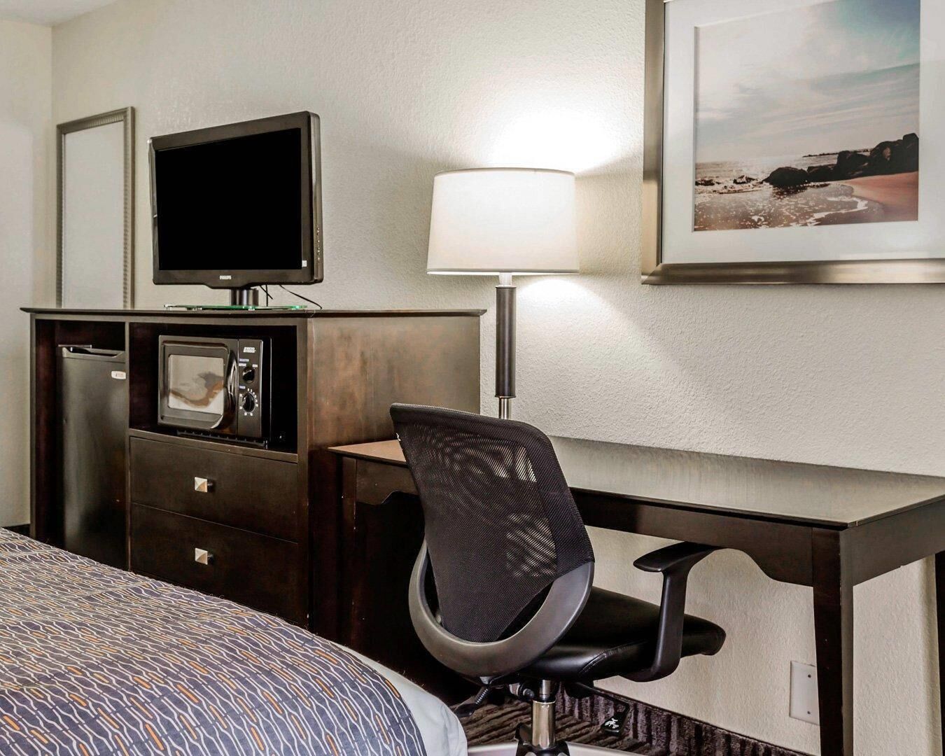 A hotel room with a bed, a desk with a chair, a television, a lamp, a refrigerator, and a framed picture on the wall.