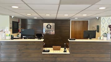 A hotel reception desk with a 'Quality by Choice Hotels' sign, two computer monitors, a basket, and a small plant.