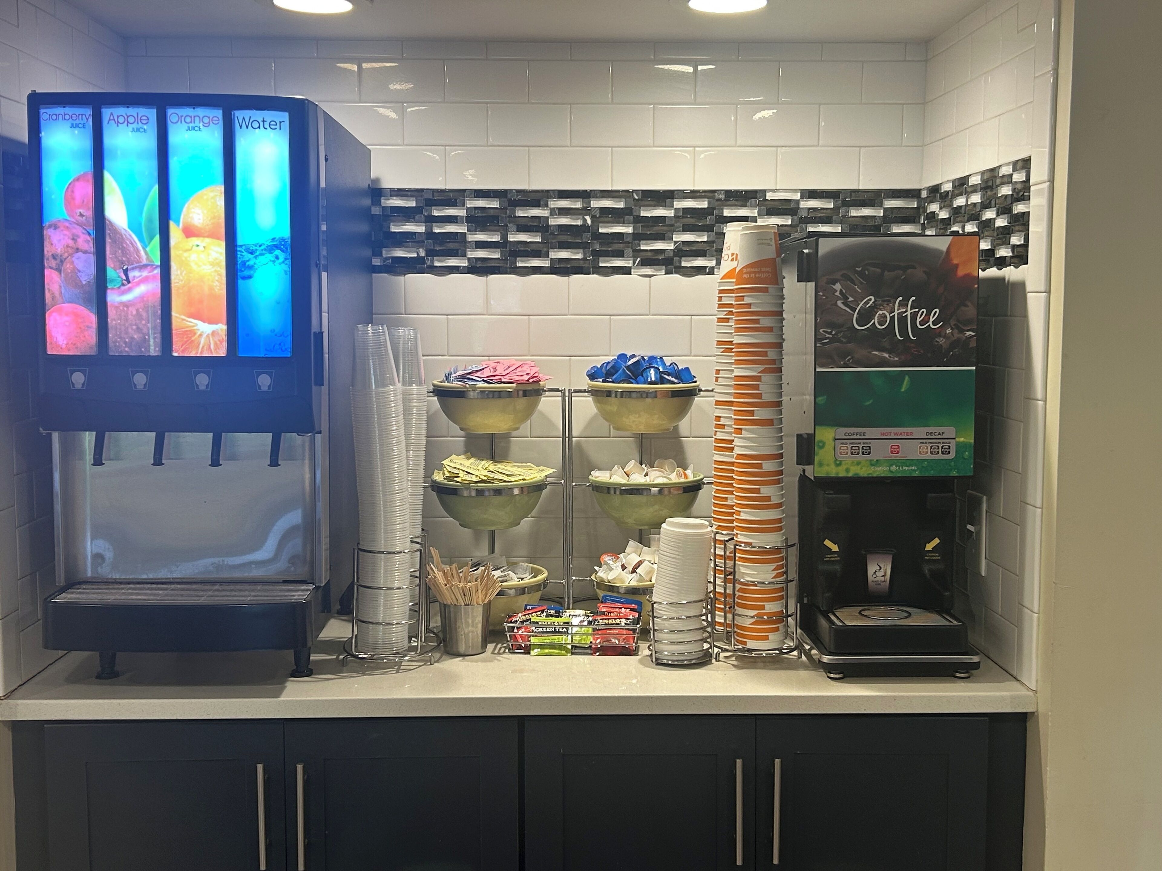 Una estación de bebidas autoservicio con opciones de jugo de arándano, manzana, naranja y agua, junto con dispensadores de café y té.
