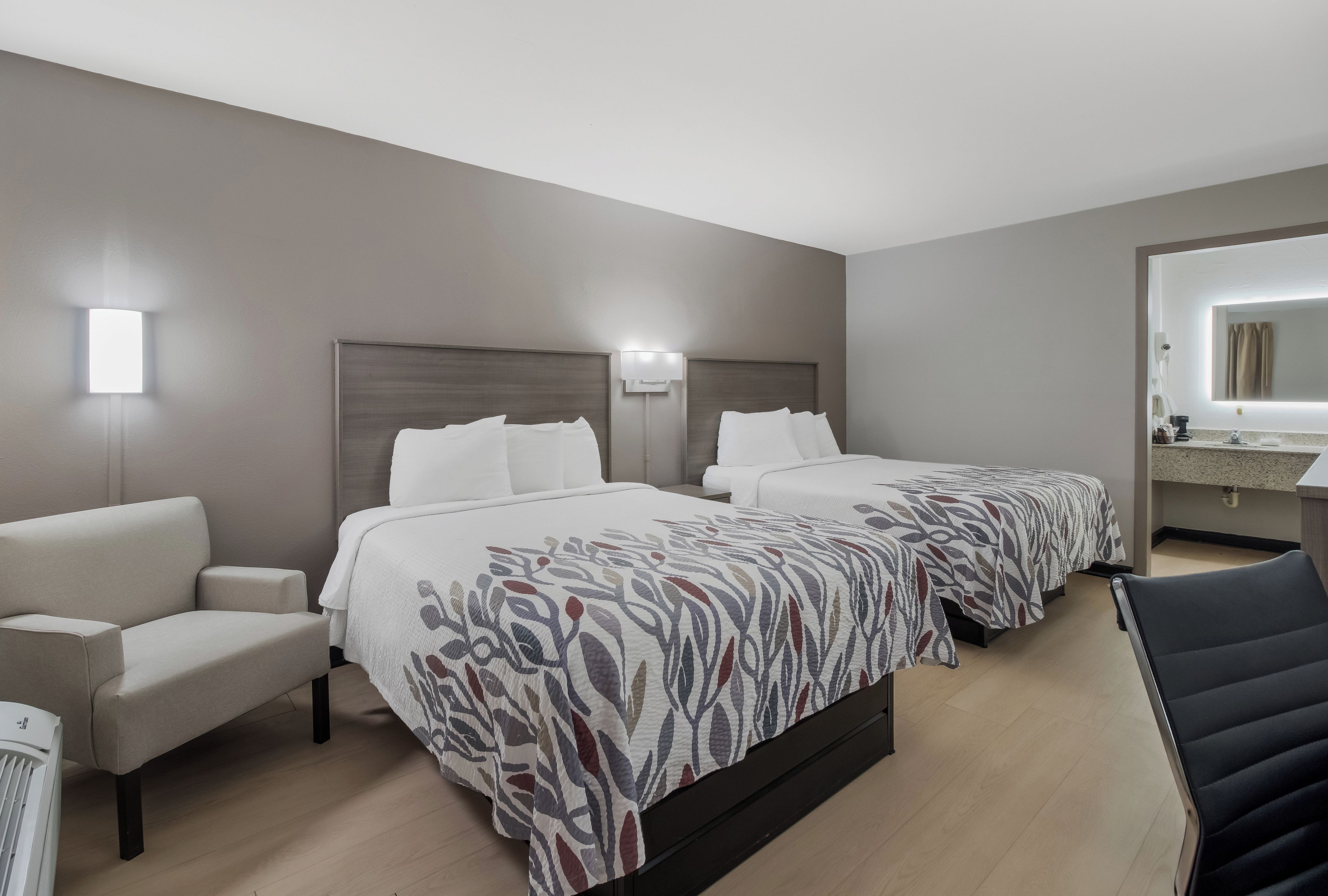A hotel room with two beds, a sofa, a chair, a desk, and a bathroom with a mirror.