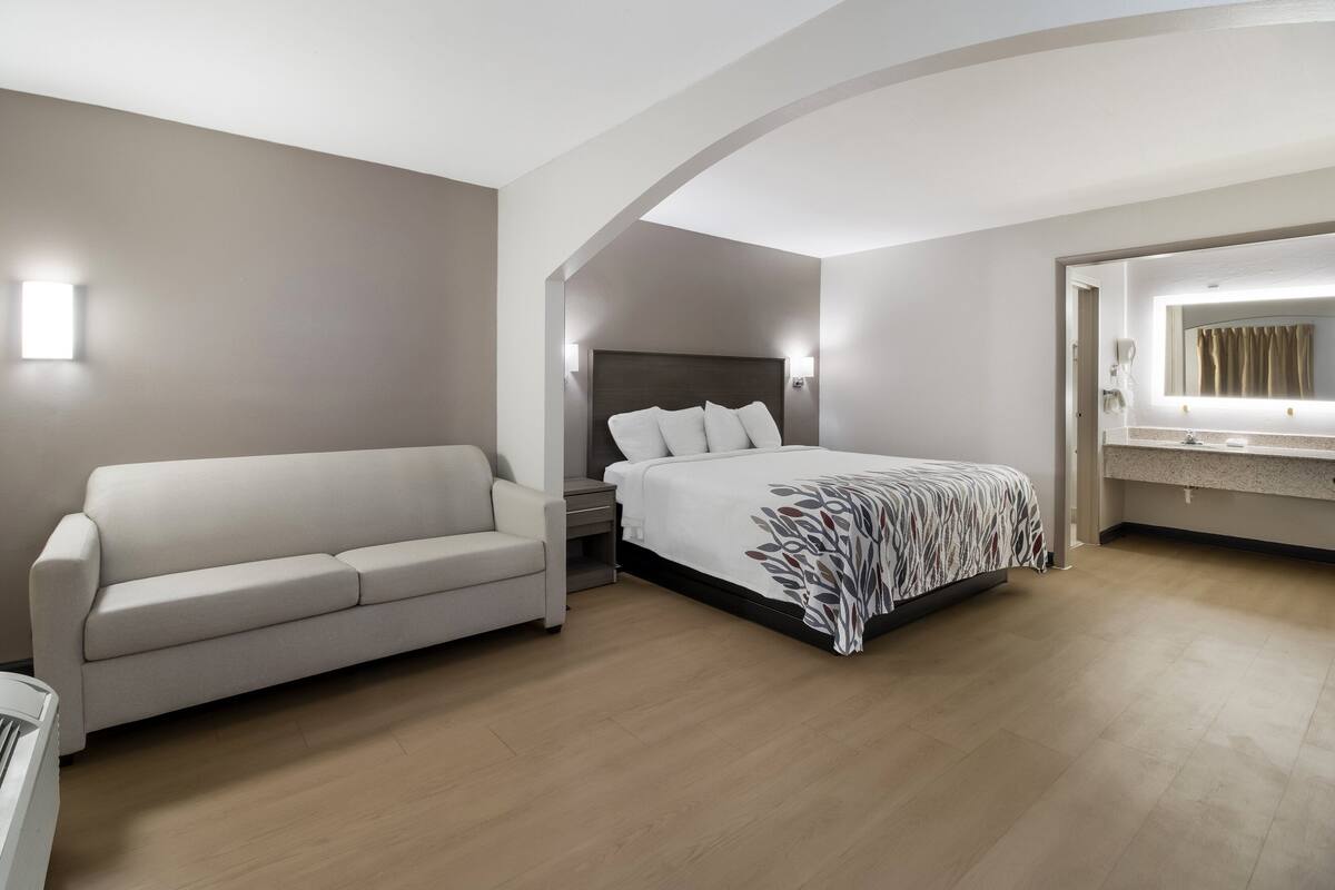 A modern hotel room with a bed, a sofa, a nightstand, and a bathroom with a mirror and sink.