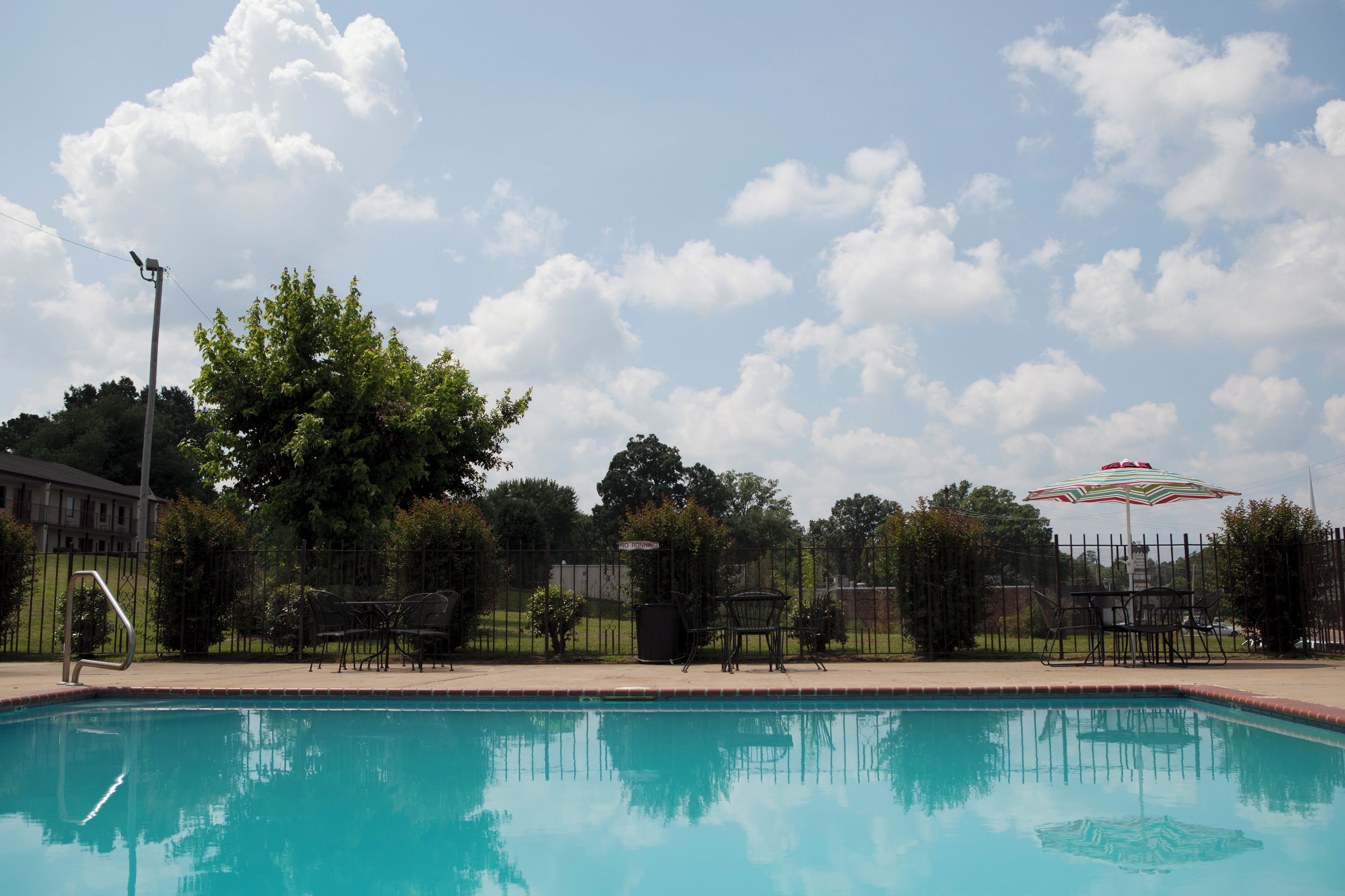 Una piscina con una silla de metal y una sombrilla, rodeada por una valla y árboles.