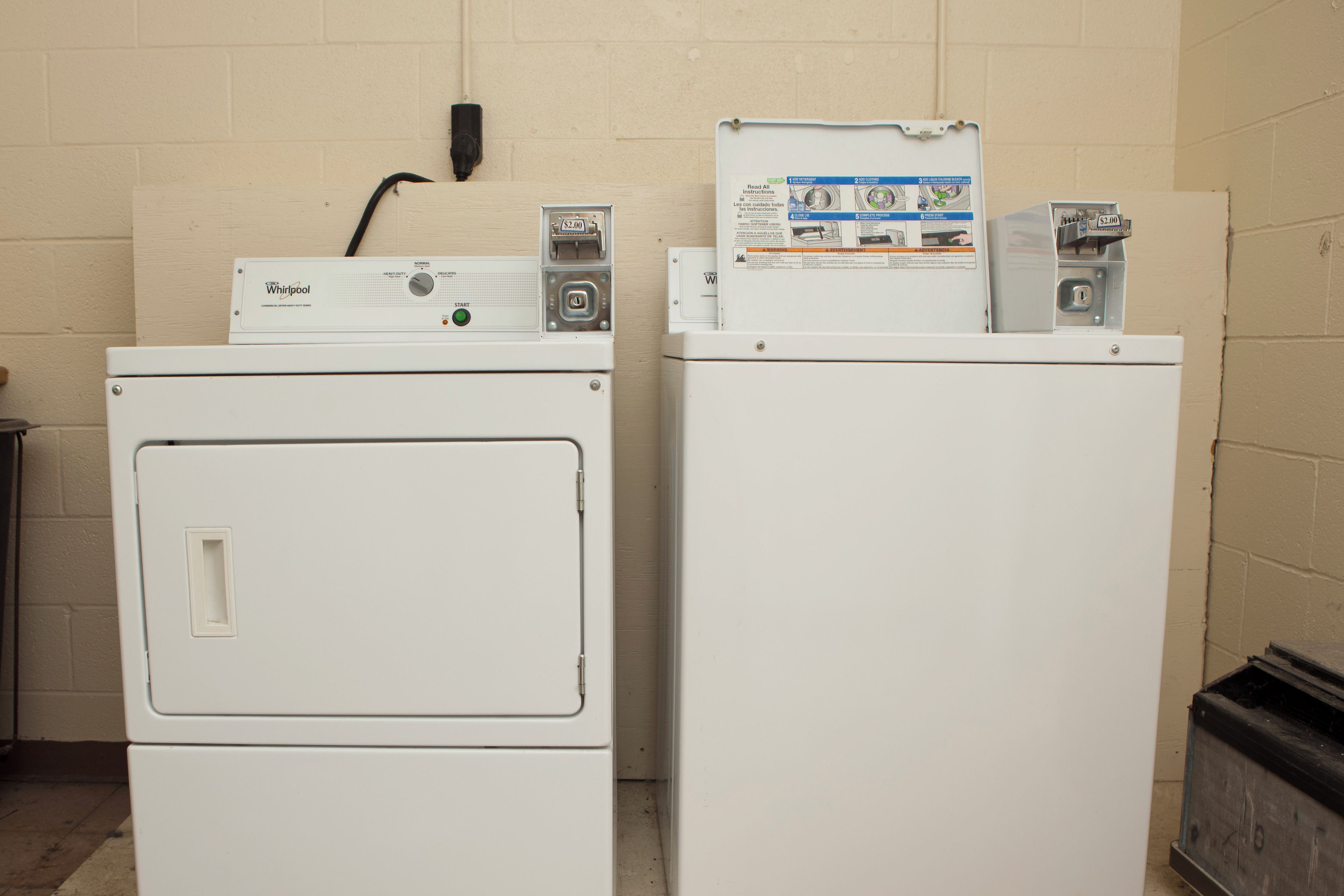 Two stacked Whirlpool front-loading washing machines in a utility room with beige walls and a black trash bin to the left.