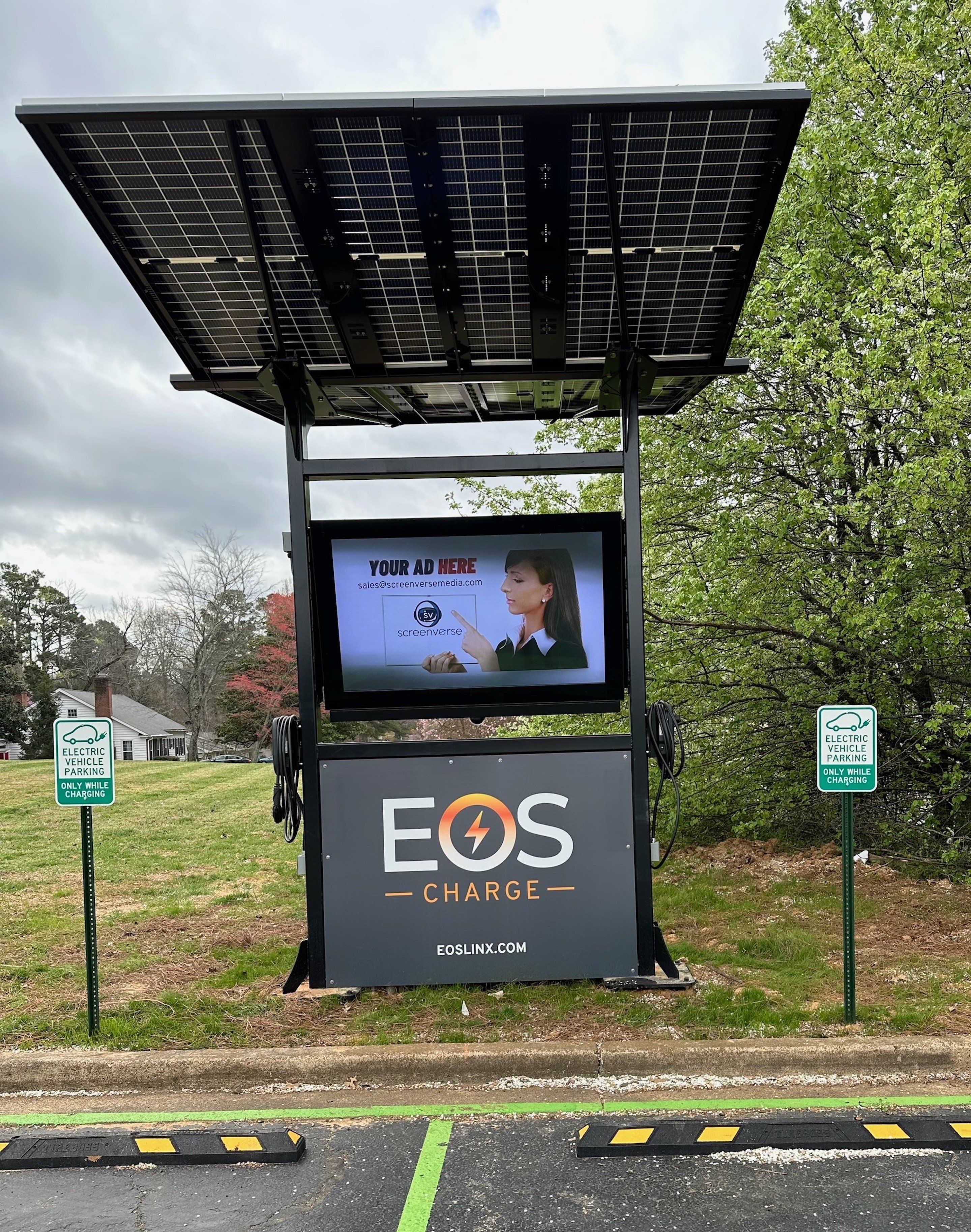 A solar-powered charging station with a screen advertising Eos Charge, surrounded by greenery and a residential area.