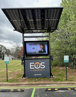 A solar-powered charging station with a screen advertising Eos Charge, surrounded by greenery and a residential area.
