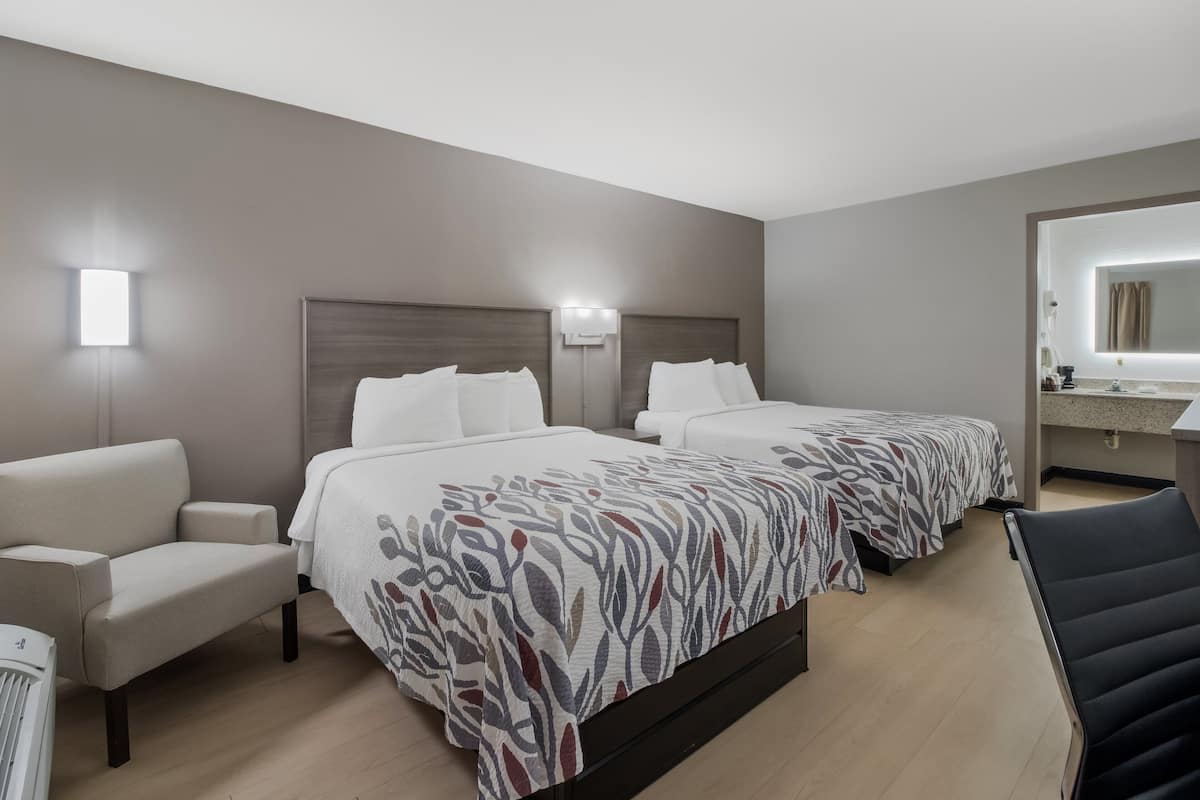 A hotel room with two beds, a sofa, a chair, a desk, and a bathroom with a mirror and sink.