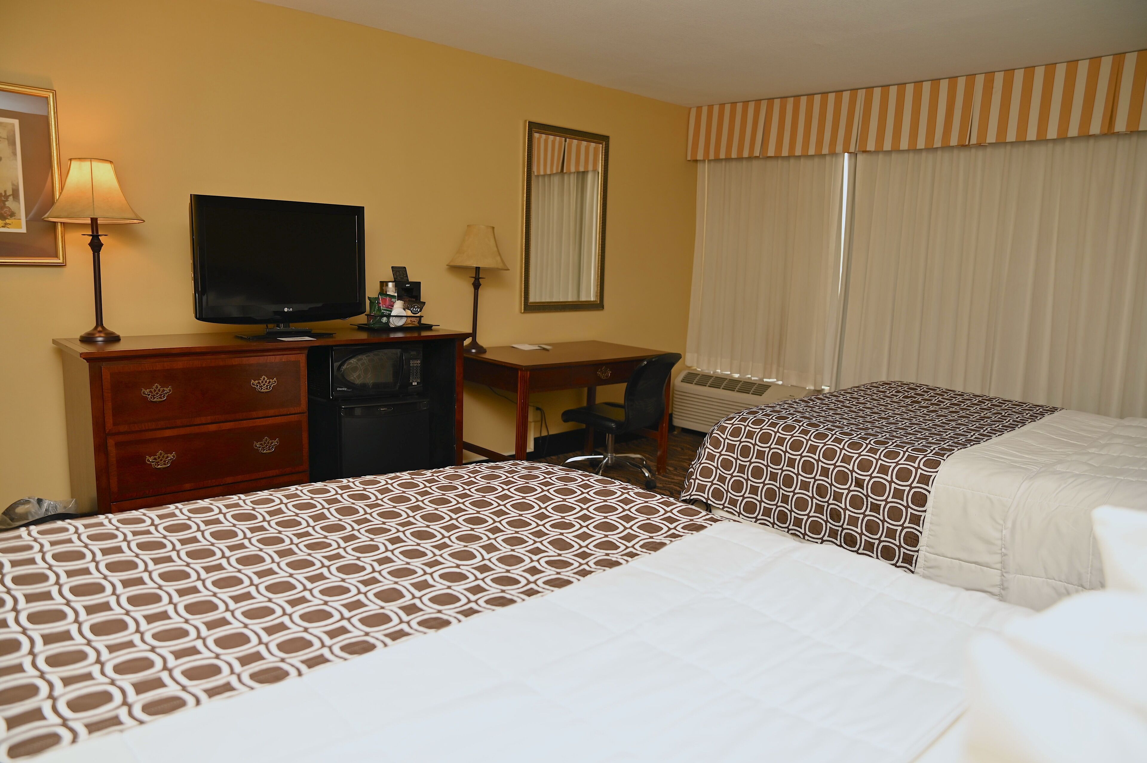 Une chambre d’hôtel avec deux lits, un bureau, une télévision, une lampe de bureau, une machine à café et une fenêtre avec des rideaux.