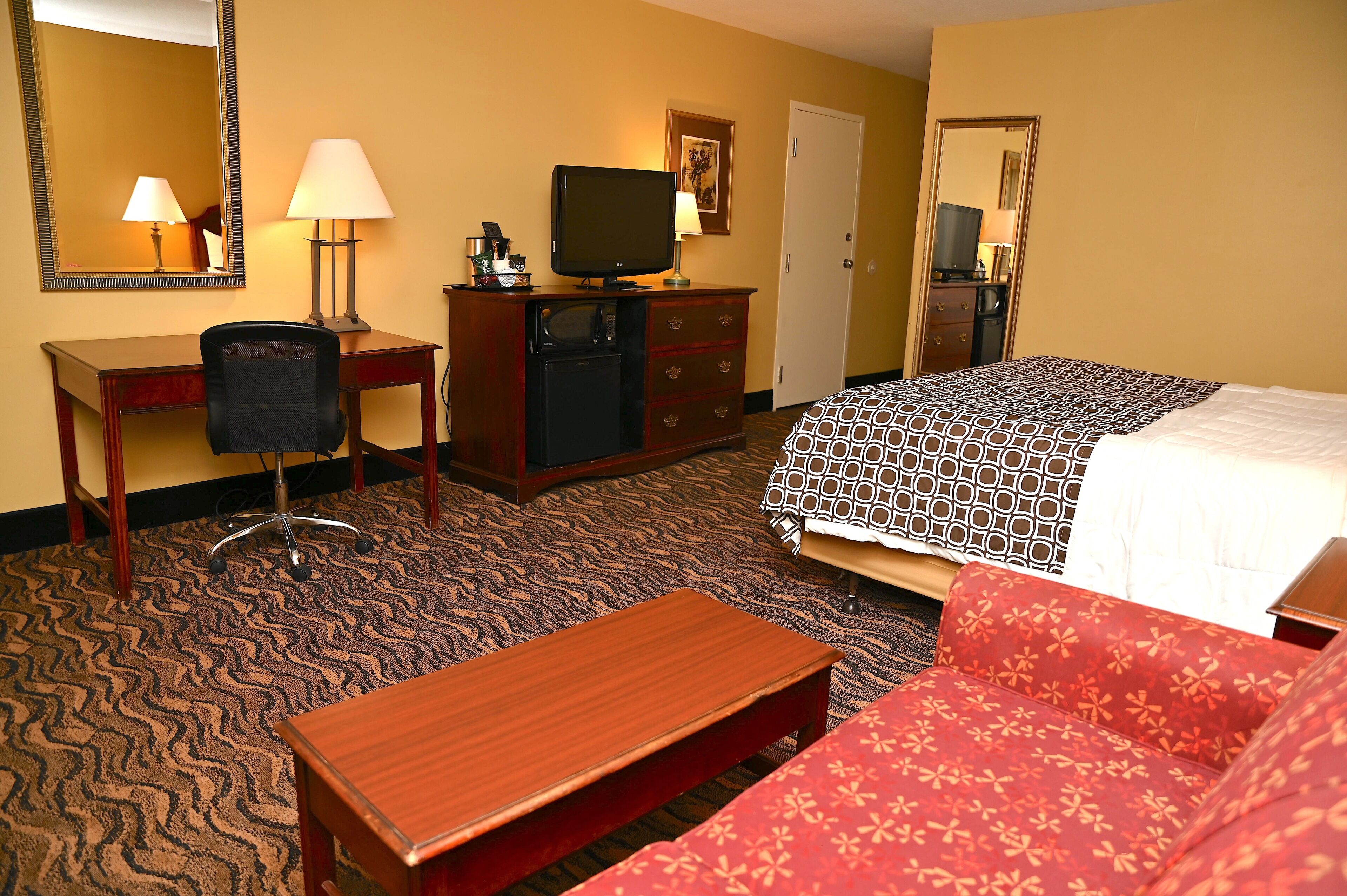 A hotel room with a bed, a desk with a chair, a television, a small refrigerator, and a bedside table.