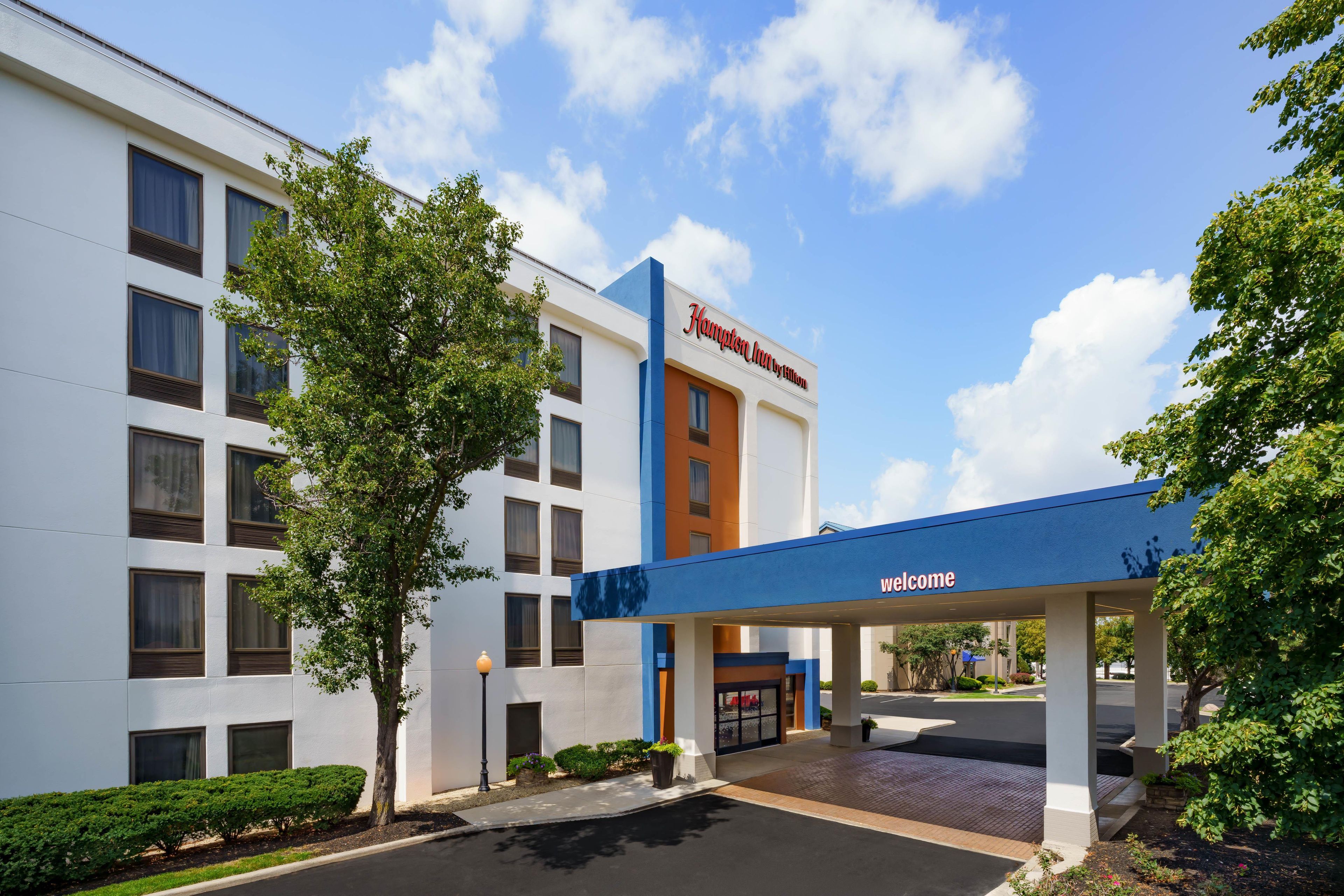 Hampton Inn by Hilton -hotelli, jossa on näkyvillä «Tervetuloa»-kyltti, parkkipaikka ja useita ikkunoita sisältävä julkisivu.