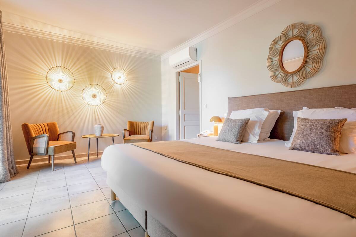 A hotel room with a large bed, two chairs, a small table, a mirror, and a ceiling light fixture.