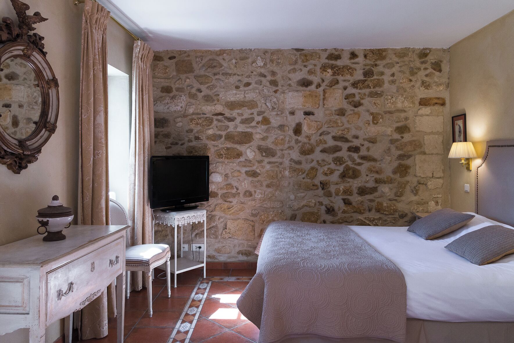 A room with a stone wall, a bed with pillows, a TV on a small table, a dresser, and a lamp.