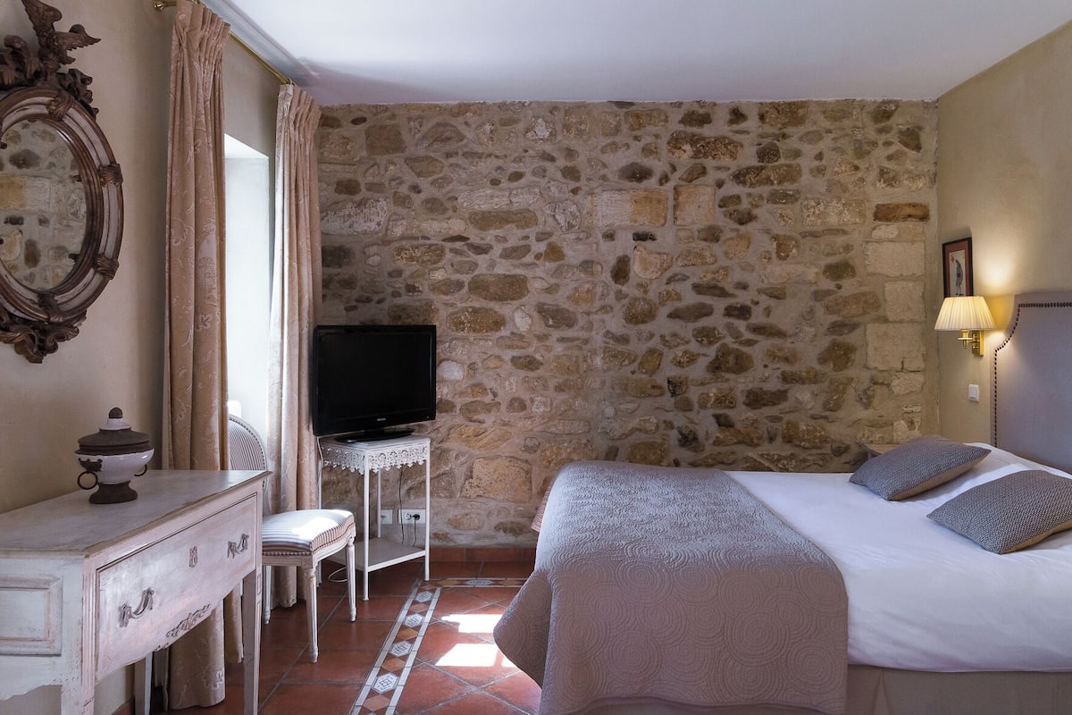 A room with a stone wall, a bed with pillows, a TV on a small table, a dresser, and a lamp.