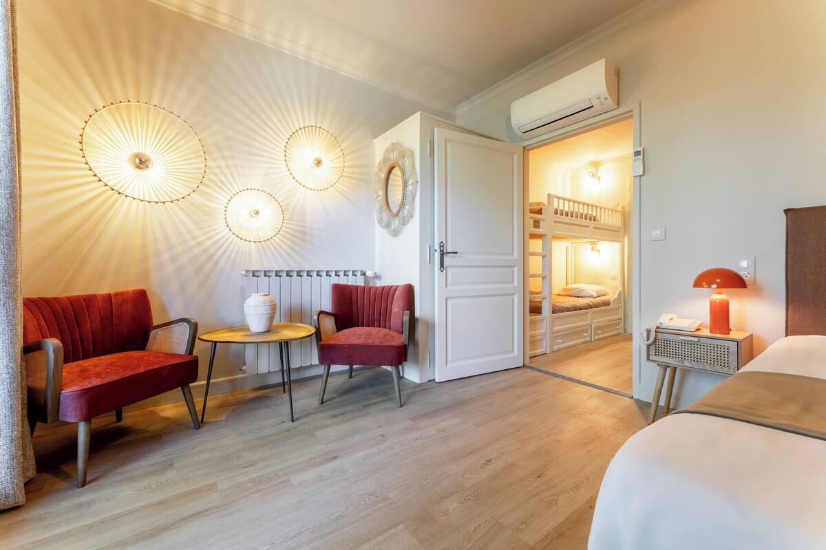 A hotel room with a bed, a red sofa, a small table, and a chair. There is a door leading to a balcony with a railing.